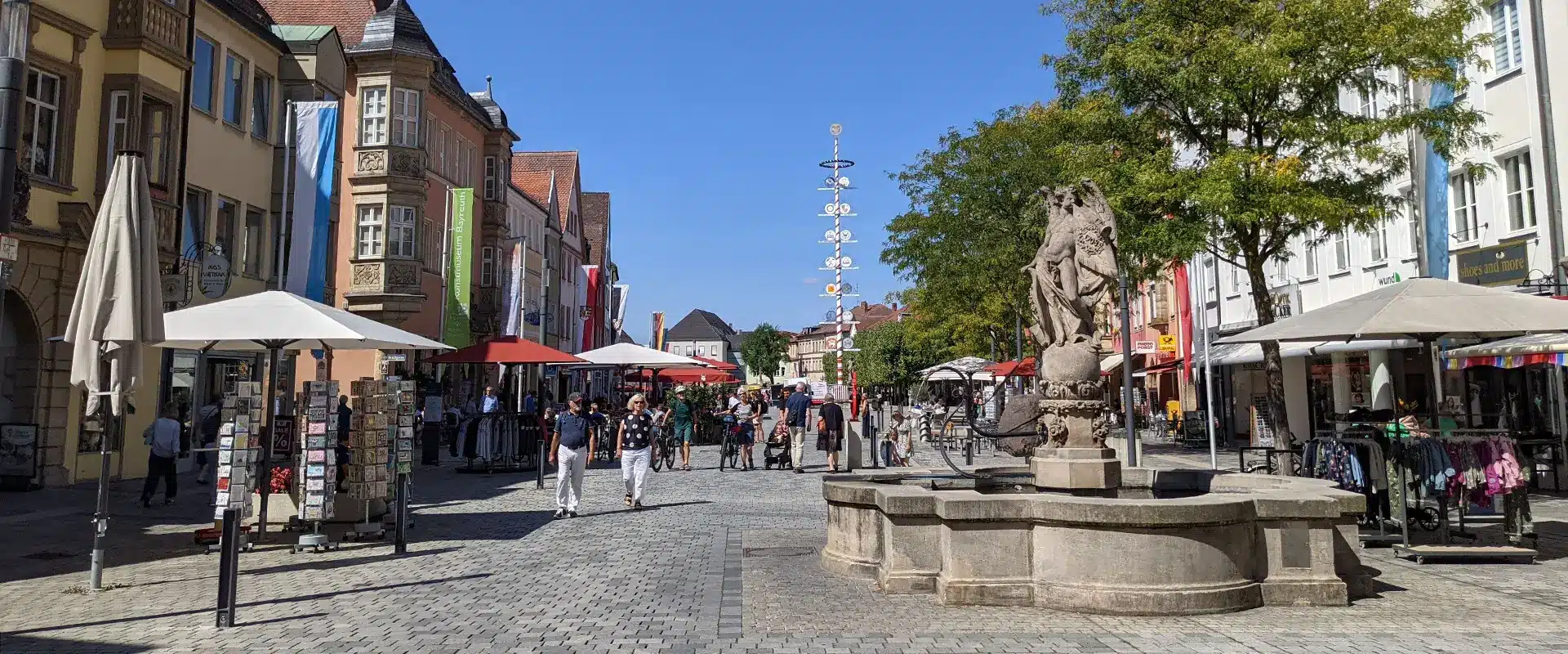 Die Innenstadt von Bayreuth mit einem Brunnen im Vordergrund, Geschäfte und einem Maibaum