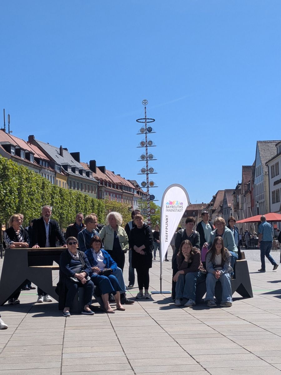 Mehrere Menschen sitzen auf den Sitztreppen, dazwischen eine Drop-Flac mit dem Innenstadtlogoe und weiter Menschen, im Hintergund der Markt mit dem Maibaum