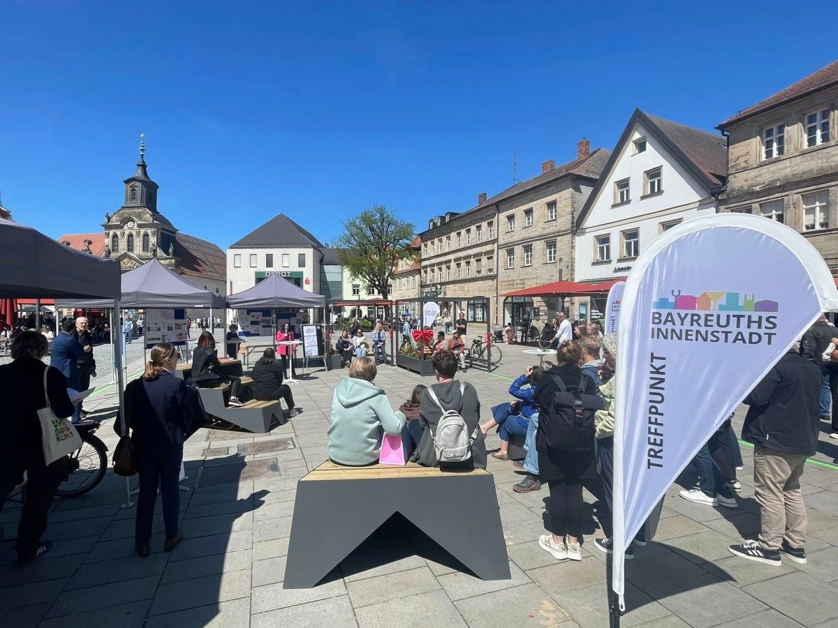 Mehrere Menschen sitzen auf den Sitztreppen, eine Dropflc mit den Innenstadtlogo und weiter Menschen stehn mit daneben und hören einer Frau (Ins Medick, if.digital) zu die an einem Stehtisch steht, im Hintergund das Stadtparkett