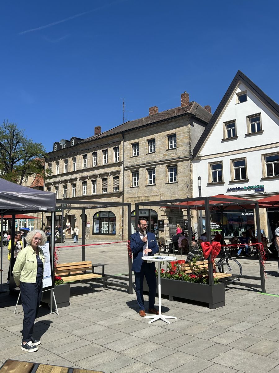 BGM A. Zippel und Baureferentin U. Kelm stehn vor den mobilen "Grünen Wohnzimmern" an eiem Stehtisch; A. Zippel hält eine Rede, im Hintergund das Stadtparkett