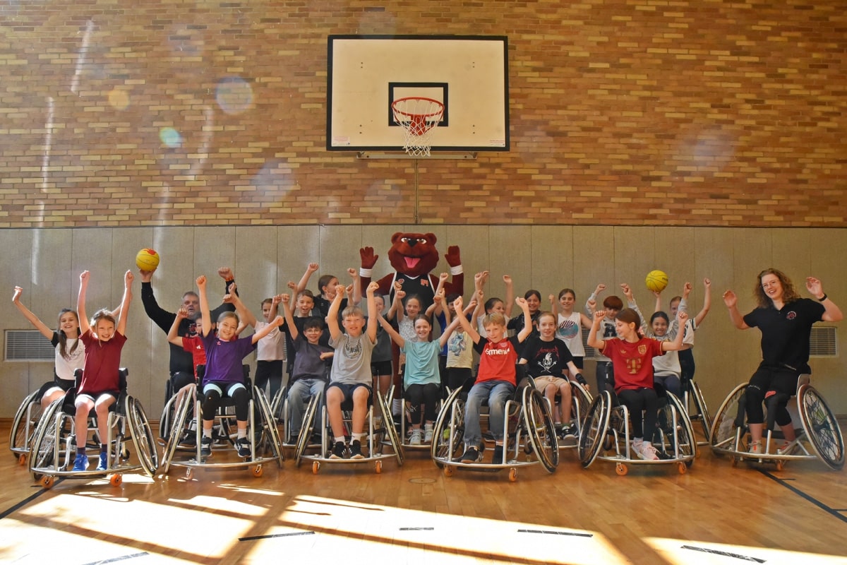 Kinder sitzen in Rollstühlen unter einem Basketballkorb und strecken ihre Arme nach oben