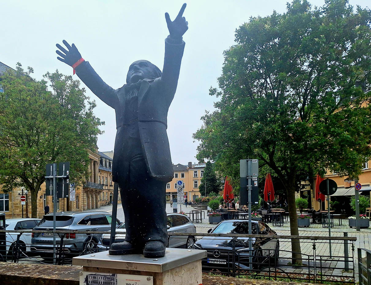 Eine Wagner-Figur steht vor parkenden Autos in einem Straßenzug. | Foto: BMTG
