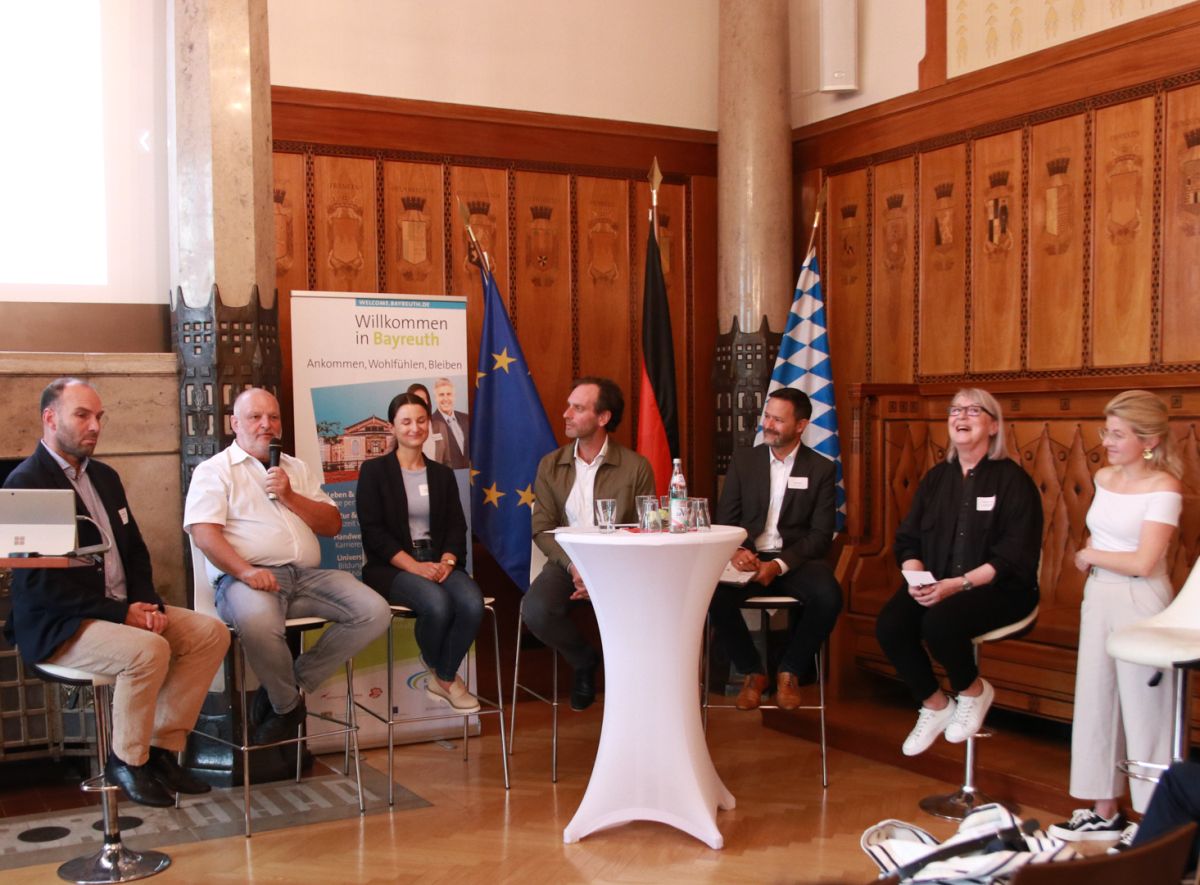 Podiumsdiskussion mit sieben Teilnehmenden auf Barhockern