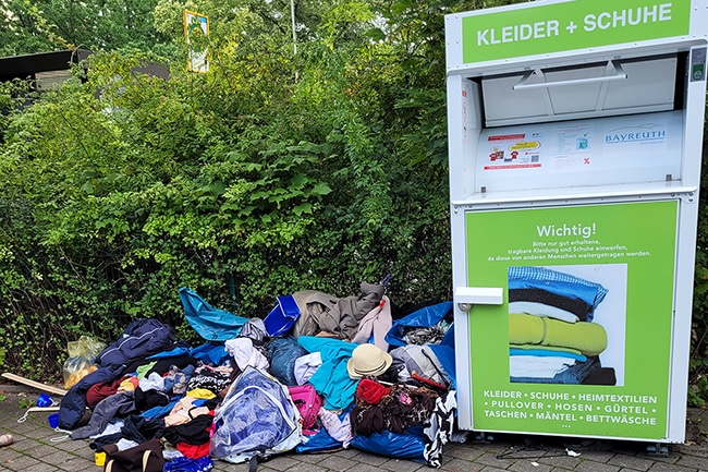 Ein Haufen Klamotten liegt zerstreut am Boden neben dem Altkleidercontainer