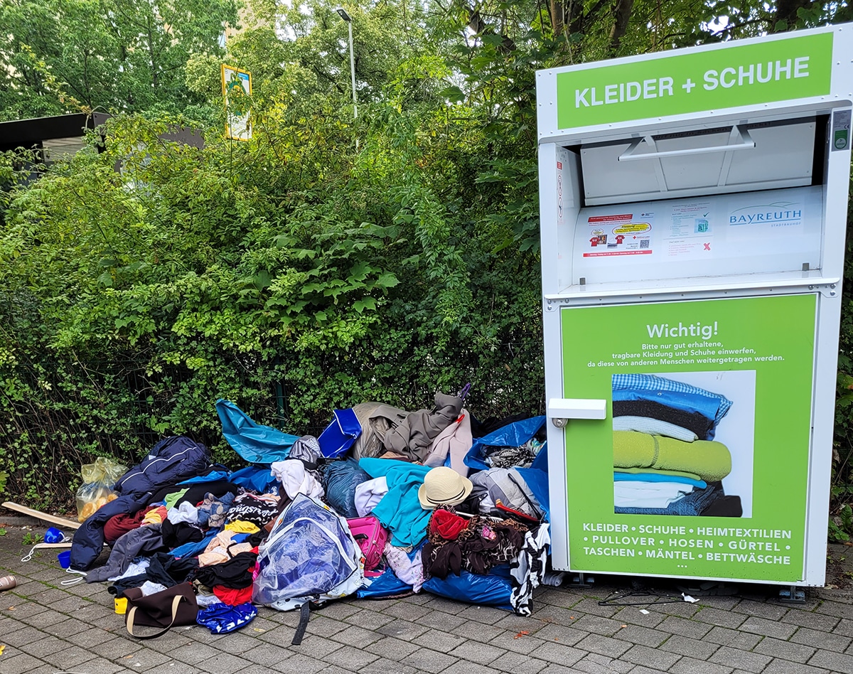 Ein Haufen Klamotten liegt zerstreut am Boden neben dem Altkleidercontainer