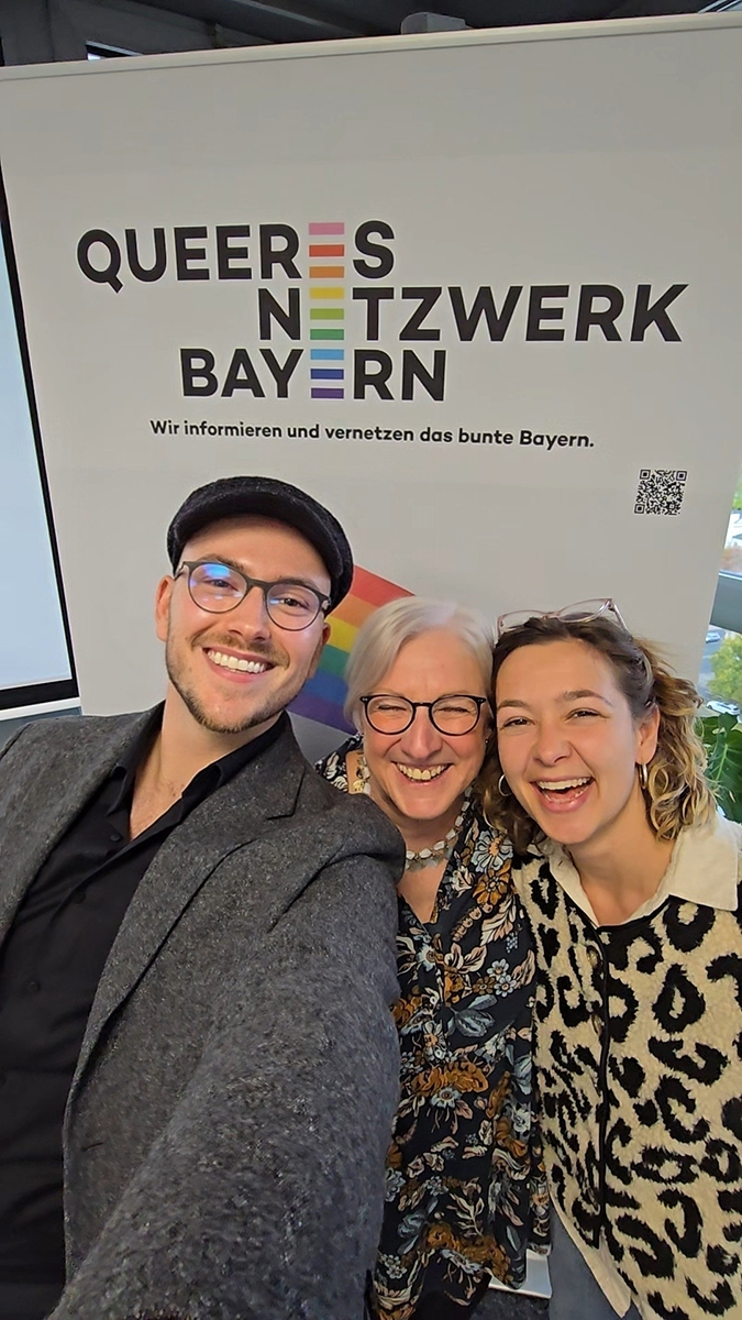 Drei lachende Menschen stehen vor einem Plakat des Queeren Netzwerk Bayern