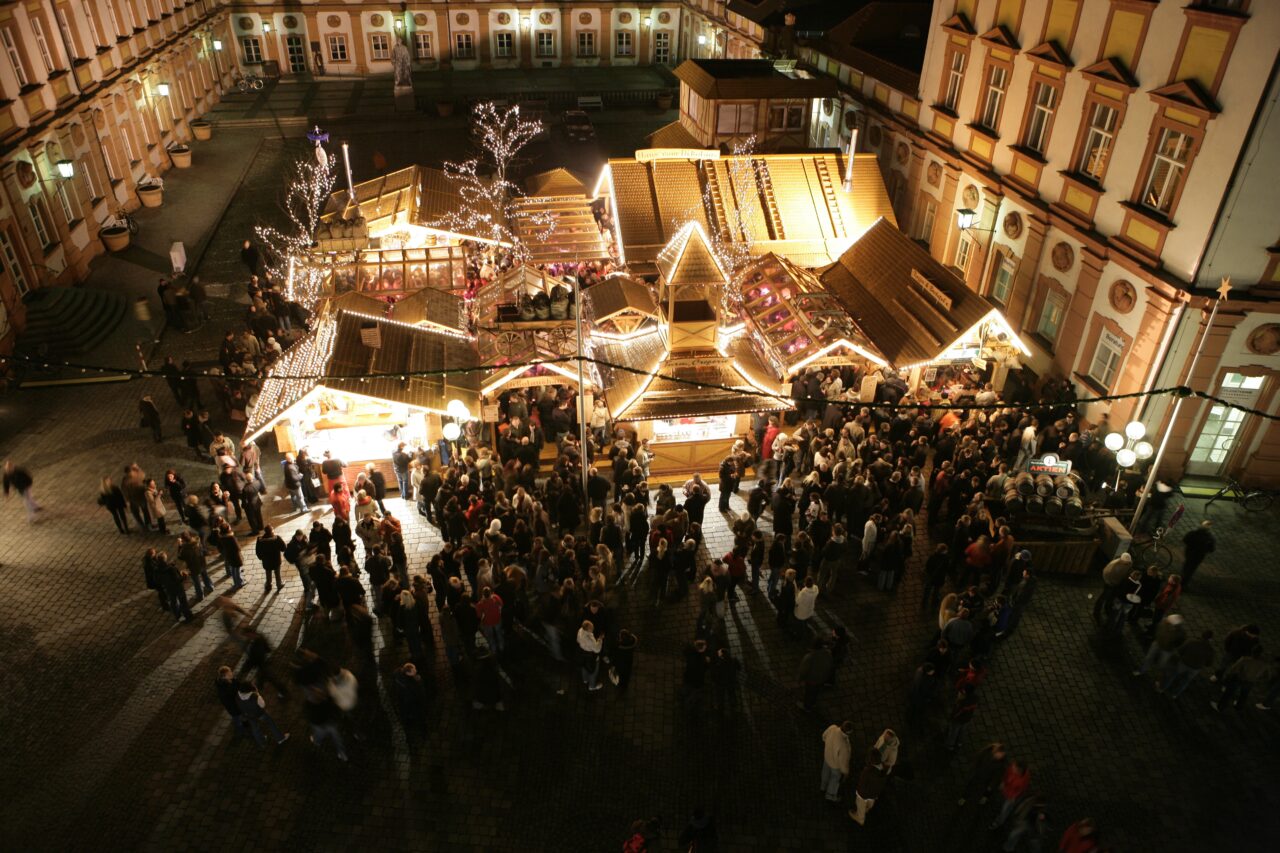 Beleuchtete Weihnachtsbuden mit Menschen an Stehtischen.