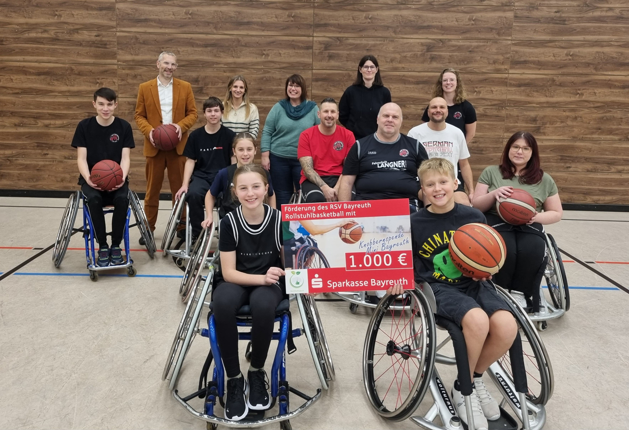 Kinder in Rollstühlen und stehende Erwachsene präsentieren in einer Turnhalle einen Spendenscheck. | Foto: Melanie Teufel / Stadtjugendring