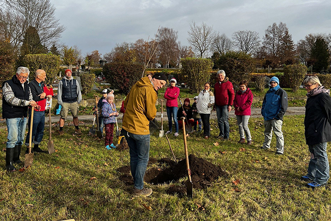 Menschen stehen auf einer Wiese, haben Schaufeln in der Hand und pflanzen einen Baum.