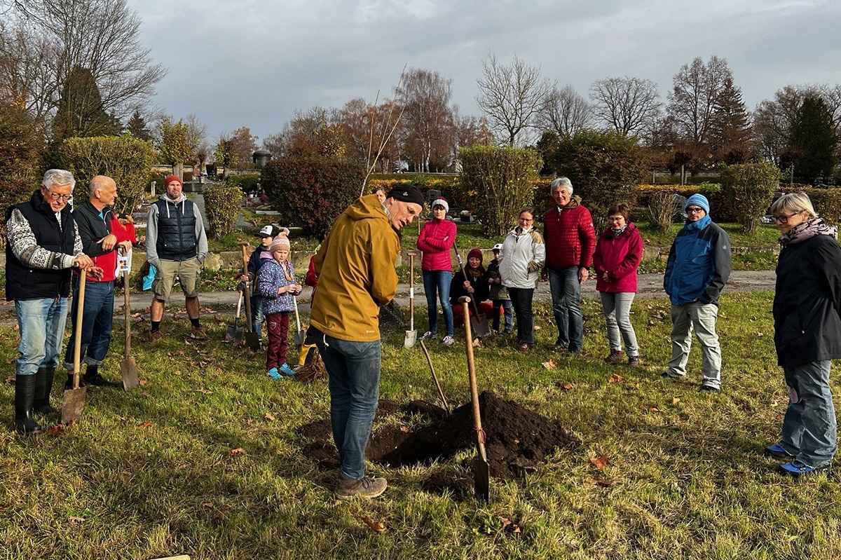 Menschen stehen auf einer Wiese, haben Schaufeln in der Hand und pflanzen einen Baum.