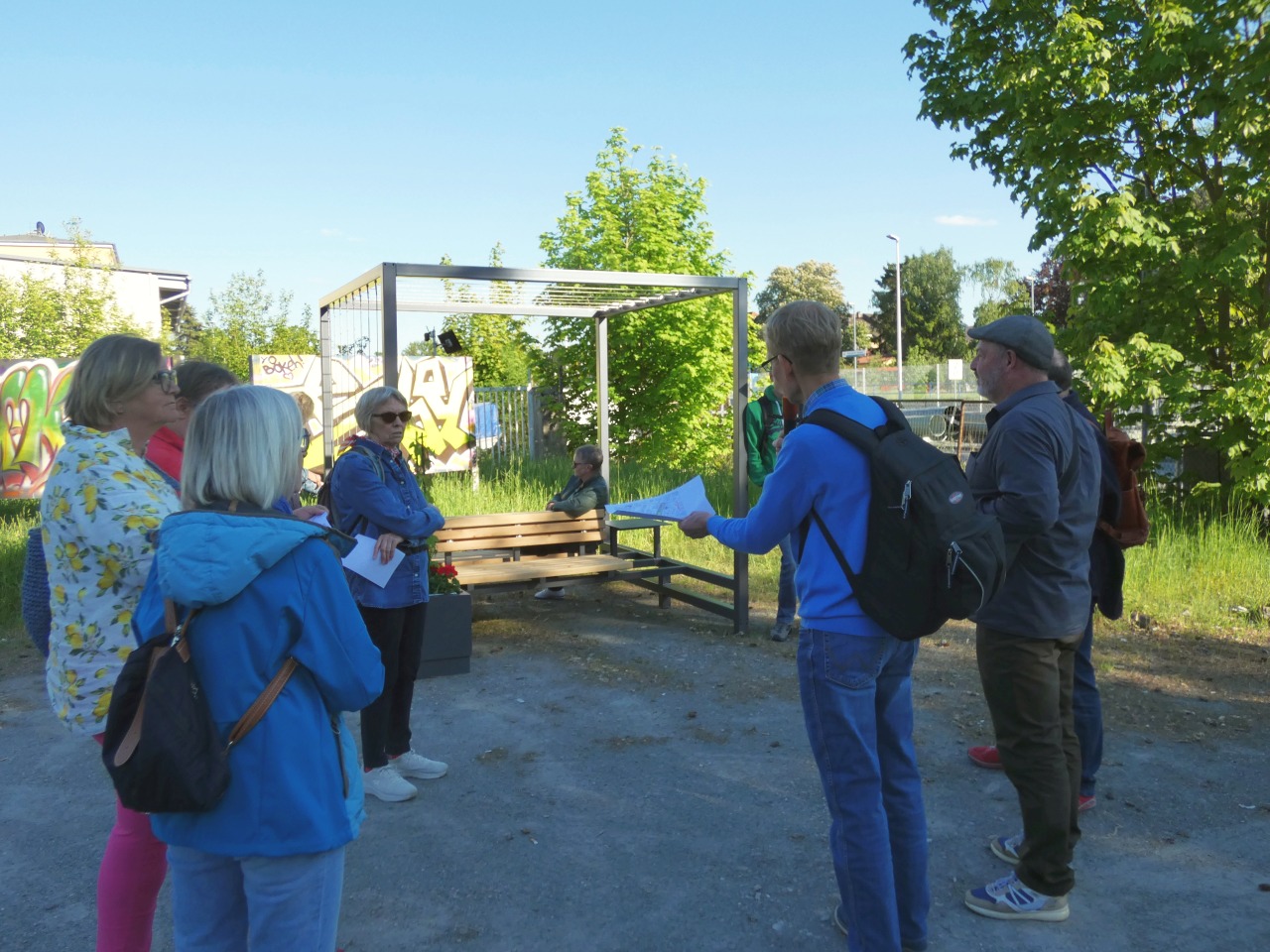 Eine kleine Gruppe von Menschen steht vor einem ortsflexiblen Sitzmöbel, aus Holzbank, Pflanztrog mit Blumen und Rankgerüst aus Stahl. Das Möbelstück soll zukünftig einen verschatteten Sitzplatz im öffentl. Raum für jedermann bieten. | Foto: Stadt Bayreuth