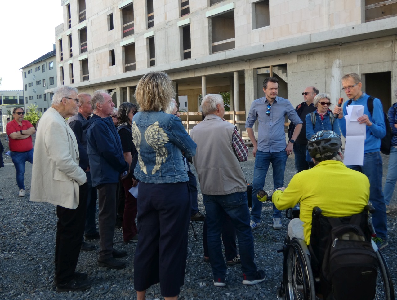 Vertreter des Tiefbauamtes und des Stadtplanungsamtes stehen vor einem Rohbaugebäude und erläutern die geplante Platzgestaltung im Stadtquartier Hugo einer Menschengruppe.