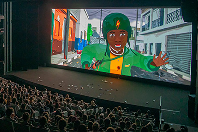 Große Leinwand auf der rechten Seite mit einem Screenshot des dort laufenden Films, davor sitzen Zuschauer