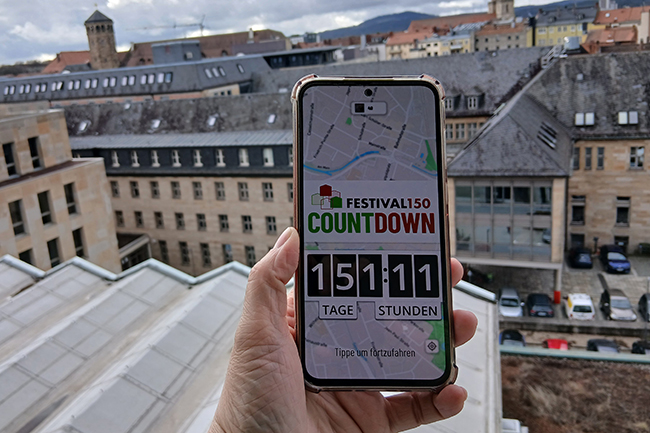 Ein Handy in der Hand, auf dem die App zu sehen ist, im Hintergrund Silhouette von Bayreuth