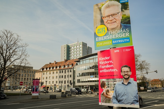 Zwei Wahlplakate zur Kommunalwahl 2026 hängen an einer Laterne. Im Hintergrund ist das Neue Rathaus zu sehen. Foto: Stadt Bayreuth