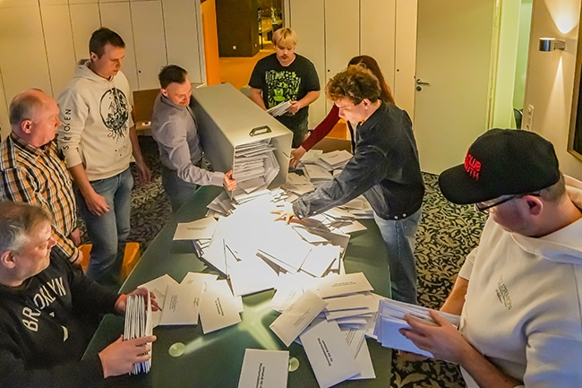 Ein Wahlhelfer entleert im Beisein von mehreren Wahlhelferinnen und -helfern eine Wahlurne auf einem Tisch. Foto: Stadt Bayreuth