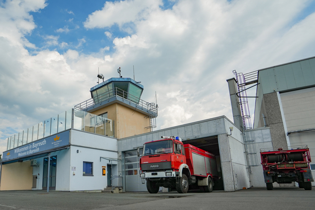 Das Hauptgebäude mit Tower des Verkehrslandeplatzes Bayreuth – hier wurden fast 13.500 Flugzeugbewegungen in 2025 gezählt. Für den Brandschutz ist mit einem speziellen Feuerwehrfahrzeug gesorgt. Die Aussichtsplattform unterhalb des Towers ist für Besucher übrigens jederzeit frei zugänglich, um den Flugbetrieb zu verfolgen. Foto: Stadt Bayreuth