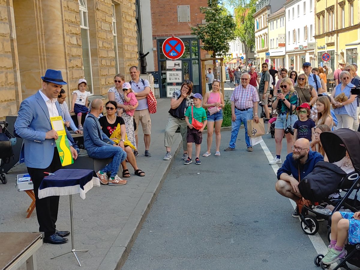 ein Mann mit Hut steht an einem kleien Zauberpult auf der Straße umringt von Publikum