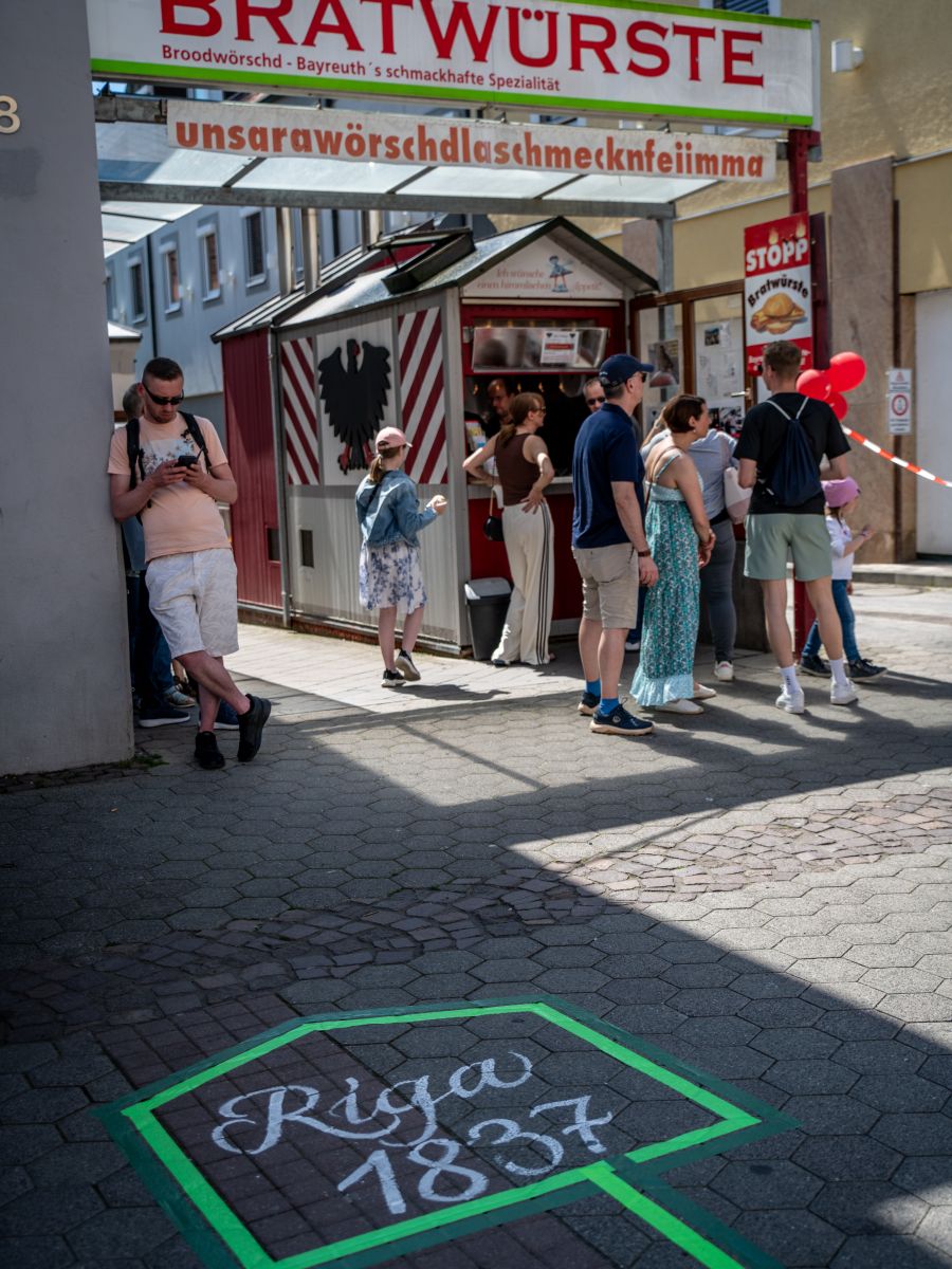 Menschen vor dem Bratwurshäuschen in der Richard-Wagner-Straße und davor ein getaptes Häuschen mit dem Schriftzug Riga 1837