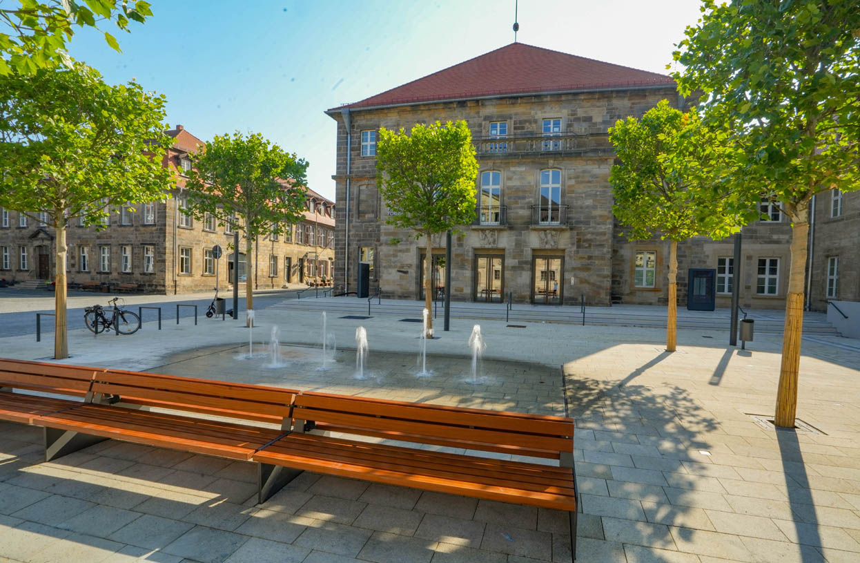 Eine große Sitzbank, mehrere Bäume und Wasserfontänen vor dem Eingang zum Friedrichsforum. | Foto: Stadt Bayreuth