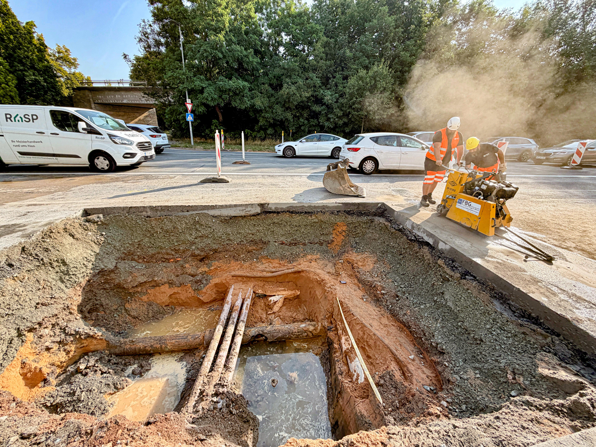 Offene Baugrube mit einem Wasserleitungsrohrbruch, anderen Leitung, zwei Bauarbeitern, die die Asphaltdecke aufschneiden und mehreren Fahrzeugen im Hintergrund. Foto: Stadt Bayreuth