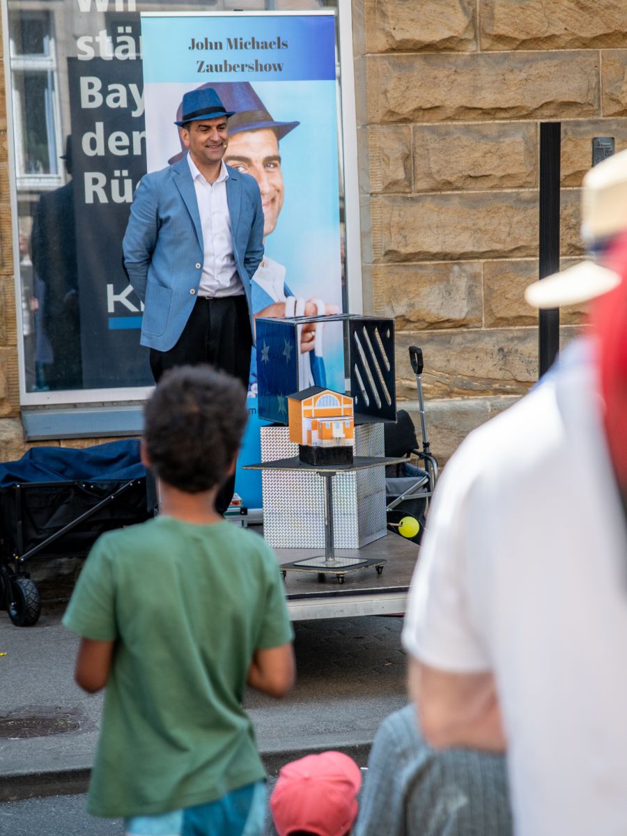 Ein Zauberer in blauen Sakko und mit blauen Hut steht auf eine Straßenbühne und enthüllt ein kleines Festspielaus, davor Publikum