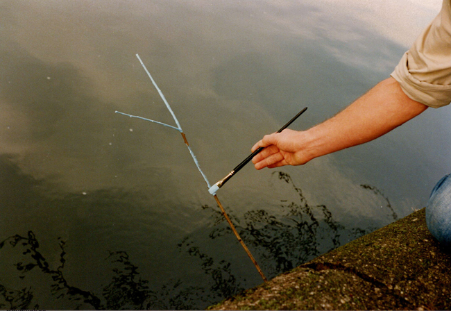 Zu sehen ist ein Ast auf einer Wasseroberfläche, der von einem Pinsel in einer Hand blau angemalt wird.